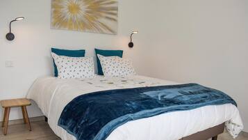 Un lit bien fait, avec une couette blanche et bleue, deux coussins turquoise et un plaid bleu. Deux lampes de lecture sont fixées au mur, de chaque côté du lit.