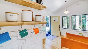 A modern kitchen with wooden shelves holding jars labeled 'SUGAR,' 'COFFEE,' and 'TEA.' A bright orange sofa and a wooden dining table are visible in the foreground. - Peaceful Mendocino Cottage Surrounded By Redwoods (Mendocino)