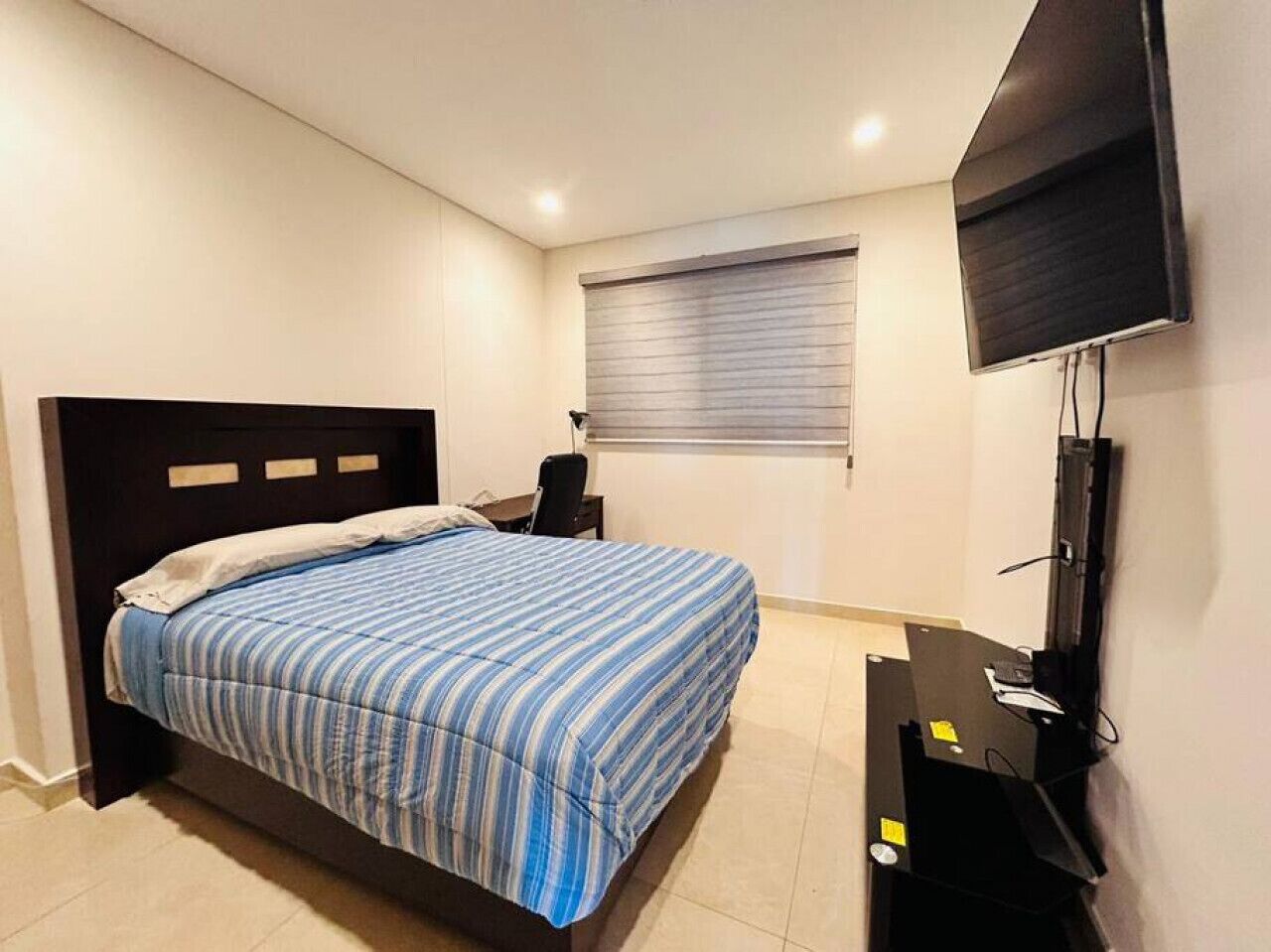 A bedroom with a bed, a desk, a chair, a television, and a window with blinds.