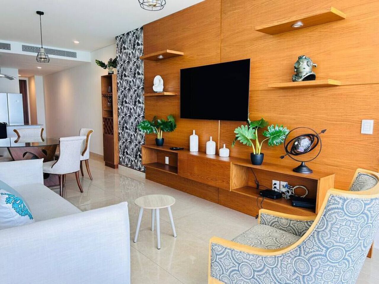A modern living room with a flat-screen TV, wooden shelving, and a dining area with white chairs.