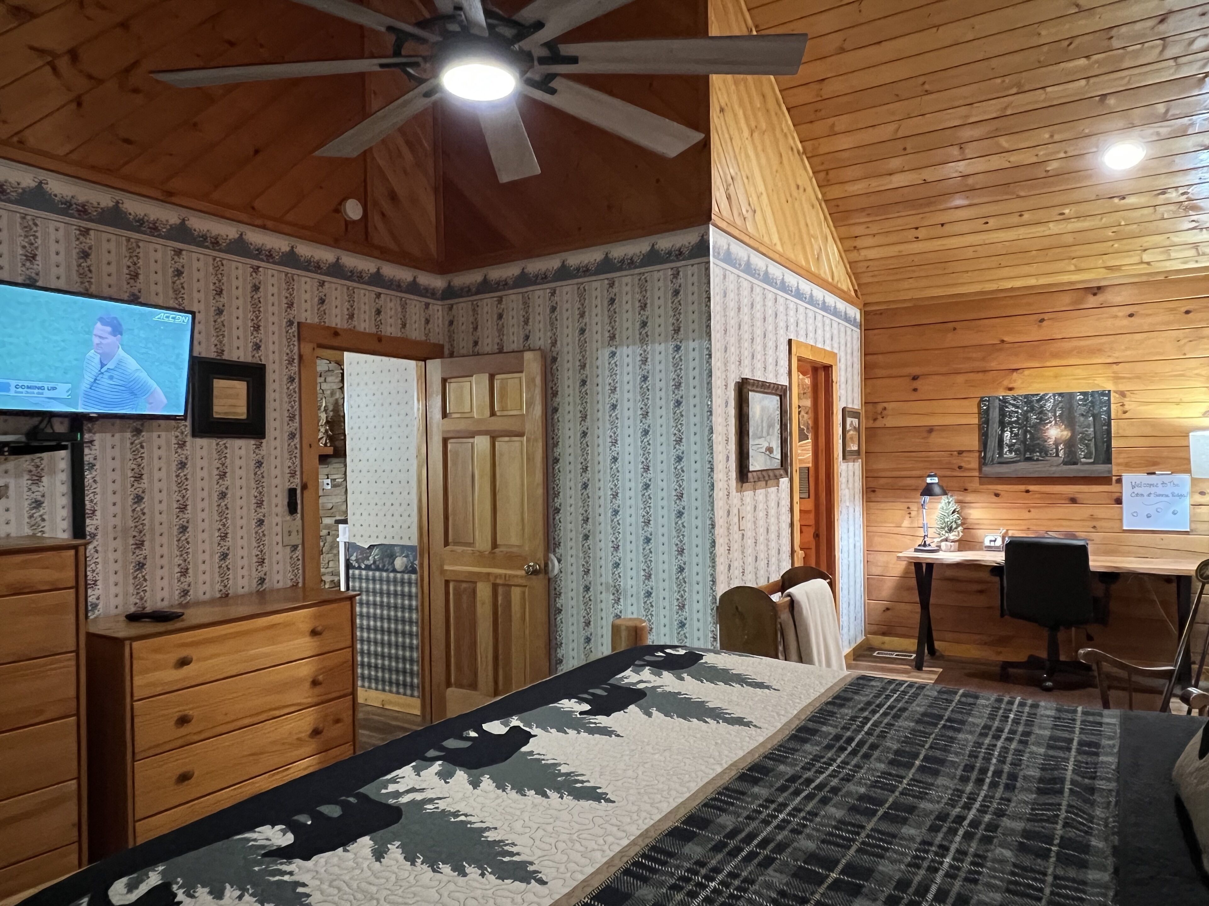 A cozy room with wooden walls, a ceiling fan, a bed with a patterned bedspread, a desk with a chair, and a television displaying a news channel.