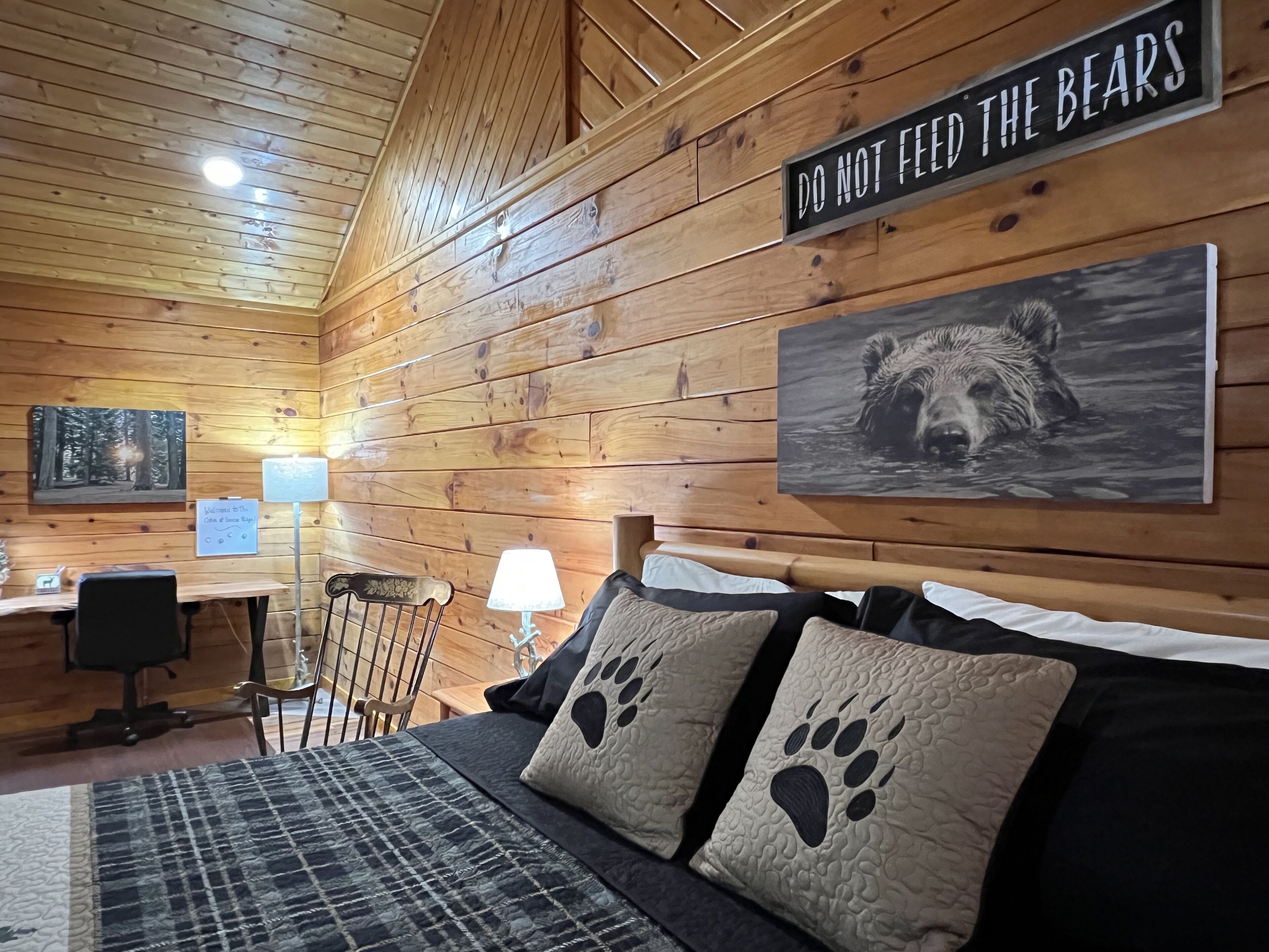 Ein gemütliches Blockhauszimmer mit Holzwänden, einem Bett mit Pfoten-Kissen, einem Schreibtisch und einem Schild mit der Aufschrift 'DO NOT FEED THE BEARS'.