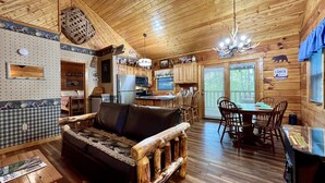 Ein Interieur einer Hütte mit Holzboden, einer Couch, einem Esstisch mit Stühlen, einer Küchenzeile und einem Deckenventilator.