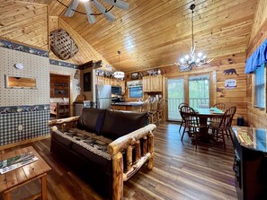 A cabin interior with a wooden floor, a sofa, a dining table with chairs, a kitchen area, and a ceiling fan. - Cozy cabin 2 miles from Parkway! Secluded_Screen Porch_Wifi_Pool_Dog Friendly (Sevierville)