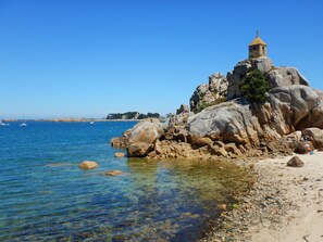 Uma área costeira rochosa com água cristalina, uma pequena torre e barcos ao longe.