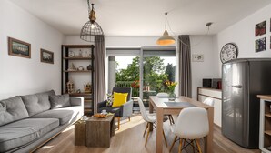 Un soggiorno moderno con un divano grigio, un tavolo da pranzo con sedie, un tavolino da caffè, un angolo cottura con frigorifero e un balcone con vista sugli alberi.