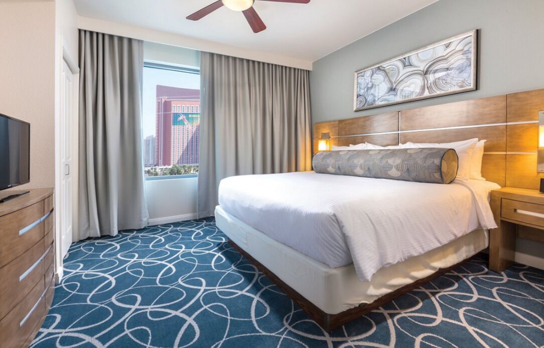 A hotel room with a large bed, a ceiling fan, a TV, a wooden dresser, and a view of a cityscape through a window with curtains.