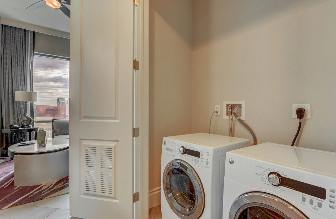 A hotel room with a washing machine, a small table with a lamp, and a view of the city through the window.