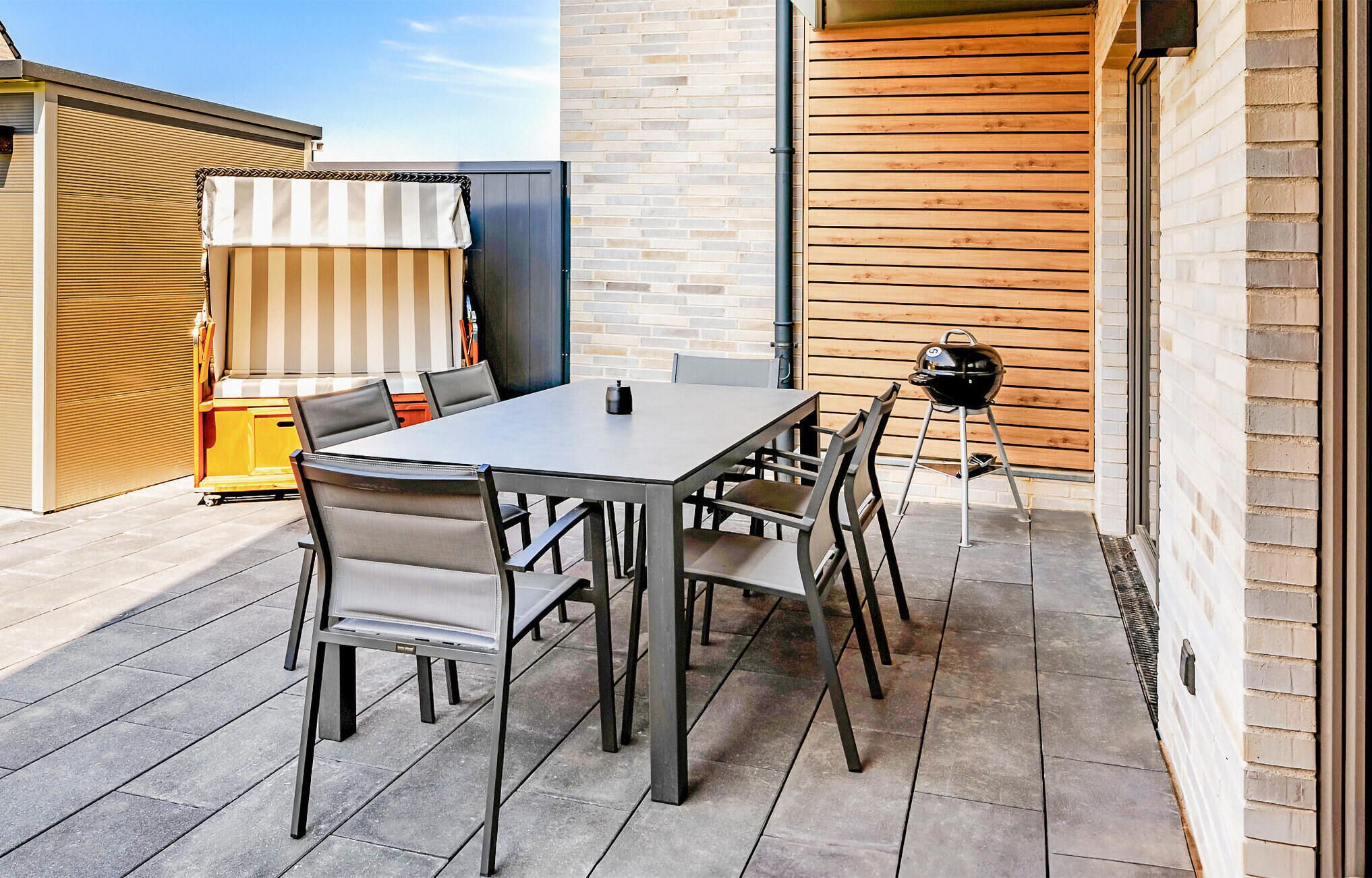 Een buitenterras met een rechthoekige tafel en vier stoelen, een zwarte barbecue en een gestreept zonnescherm.
