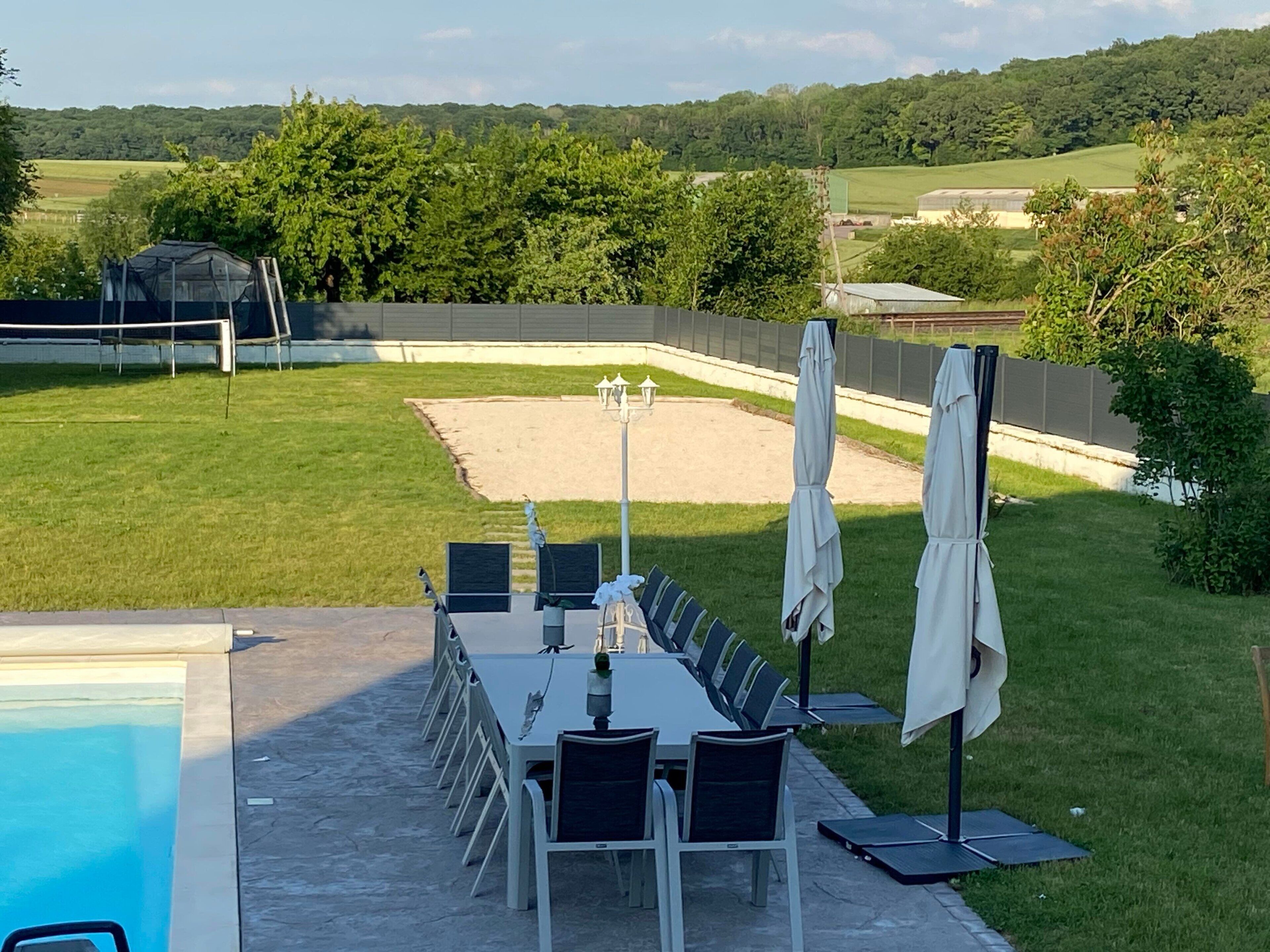 A patio with a pool, outdoor seating, and a view of a green field and trees.