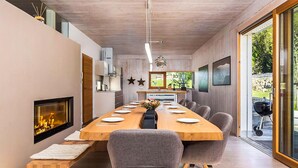Una sala da pranzo moderna con un lungo tavolo di legno, sedie grigie rivestite in tessuto, un camino e vista sull'esterno.