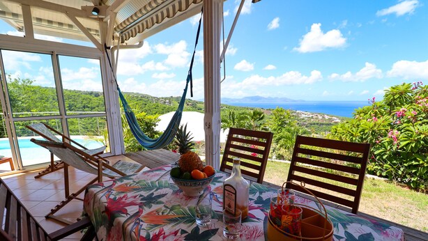 A patio with a table set for dining, a hammock, and a view of the ocean. - Villa Frangipanier with private pool (Case-Pilote)