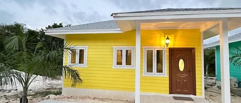 A yellow house with a brown door, a porch, and a small tree in the yard.