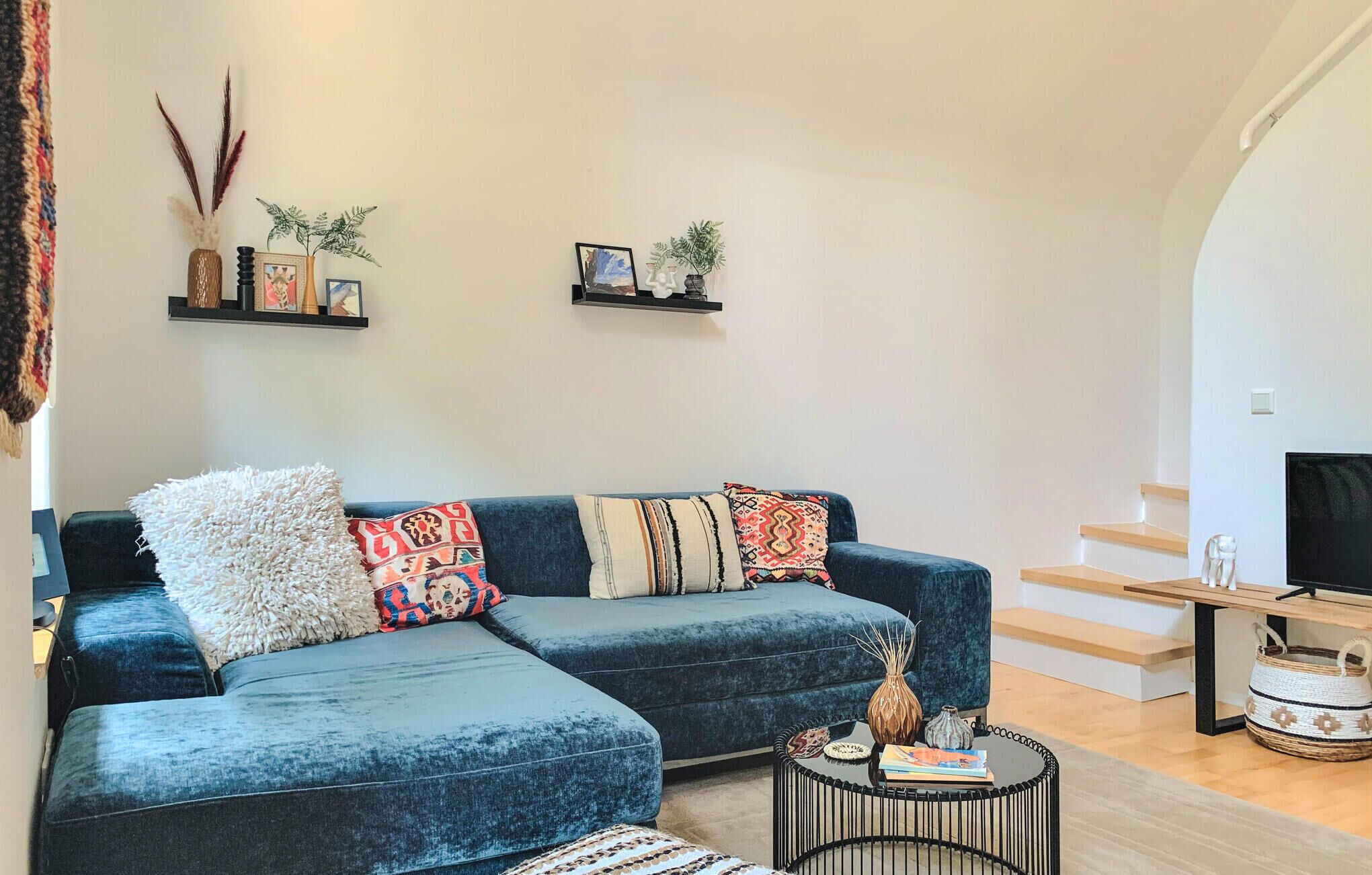 Ein modernes Wohnzimmer mit einem dunkelblauen Ecksofa, mehreren gemusterten Zierkissen, einem kleinen runden Couchtisch mit Büchern und Dekorationsobjekten sowie zwei Wandregalen mit gerahmten Bildern und kleinen Pflanzen.