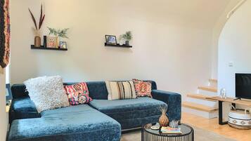 Ein modernes Wohnzimmer mit einem dunkelblauen Ecksofa, mehreren gemusterten Zierkissen, einem kleinen runden Couchtisch mit Büchern und Dekorationsobjekten sowie zwei Wandregalen mit gerahmten Bildern und kleinen Pflanzen.