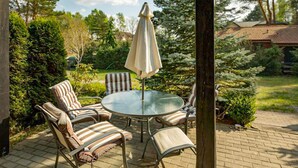 Eine Terrasse mit einem runden Glastisch und sechs gestreiften Stühlen, umgeben von Grünpflanzen und einem Pflasterweg.