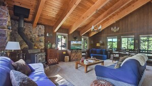 A cozy living room with a stone fireplace, wooden beams, a television, and a large window overlooking the outdoors.