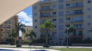 A swimming pool with a tiled deck, surrounded by palm trees and a multi-story building with balconies. - Modern Studio in Crisfer Monthly Stays WiFi & AC (Punta Cana)