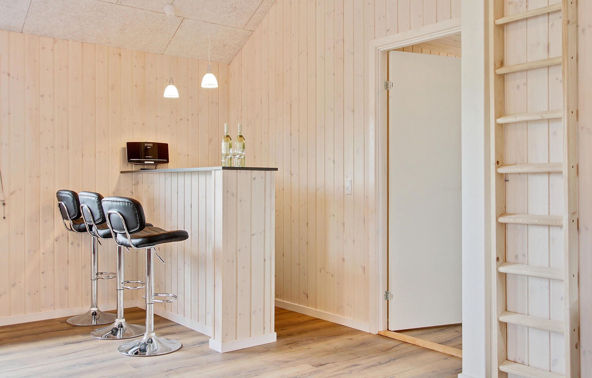 A room with wooden walls and a white door, featuring two bar stools and a small bar area with a television and bottles.