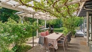 Un patio cubierto con una mesa y sillas, rodeado de follaje y un jardín.