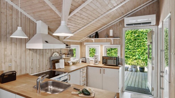 Une cuisine moderne avec des poutres en bois, des suspensions blanches, un four encastré et une vue sur un jardin luxuriant, visible à travers une porte vitrée.