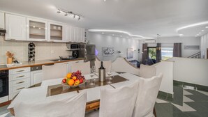 Una cocina moderna con armarios blancos, un comedor con mesa preparada para cuatro comensales y vista al mar a través de la ventana.