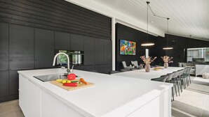 Una cocina moderna con encimera blanca, un lavamanos y una tabla de cortar de madera con verduras. Hay una zona de comedor con mesa y sillas blancas, y un cuadro abstracto de colores en la pared.