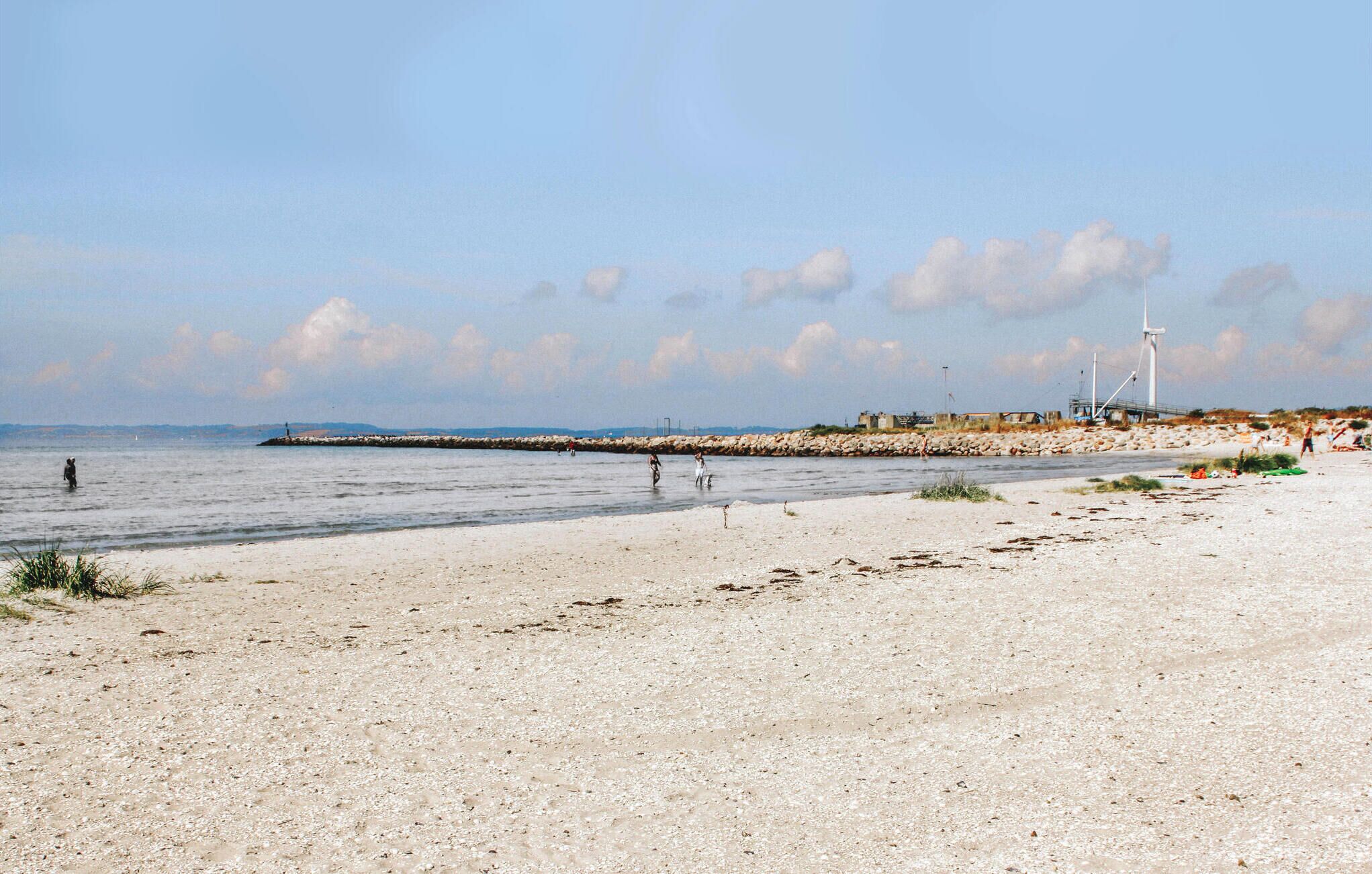 A beach with a rocky breakwater, people walking along the shore, and a lighthouse in the background.