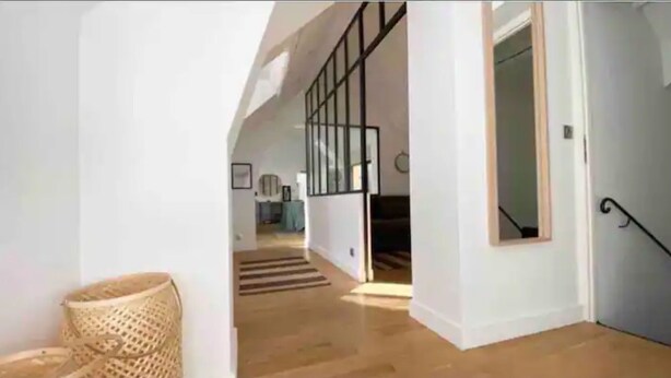 A modern interior with a staircase leading to an upper level, a wicker chair, and a kitchen area in the background. - The heart of Fontainebleau (Fontainebleau)