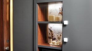 Un interior moderno con un mueble alto gris oscuro, repisas de madera y objetos decorativos, incluyendo jarrones y una superficie similar al mármol.