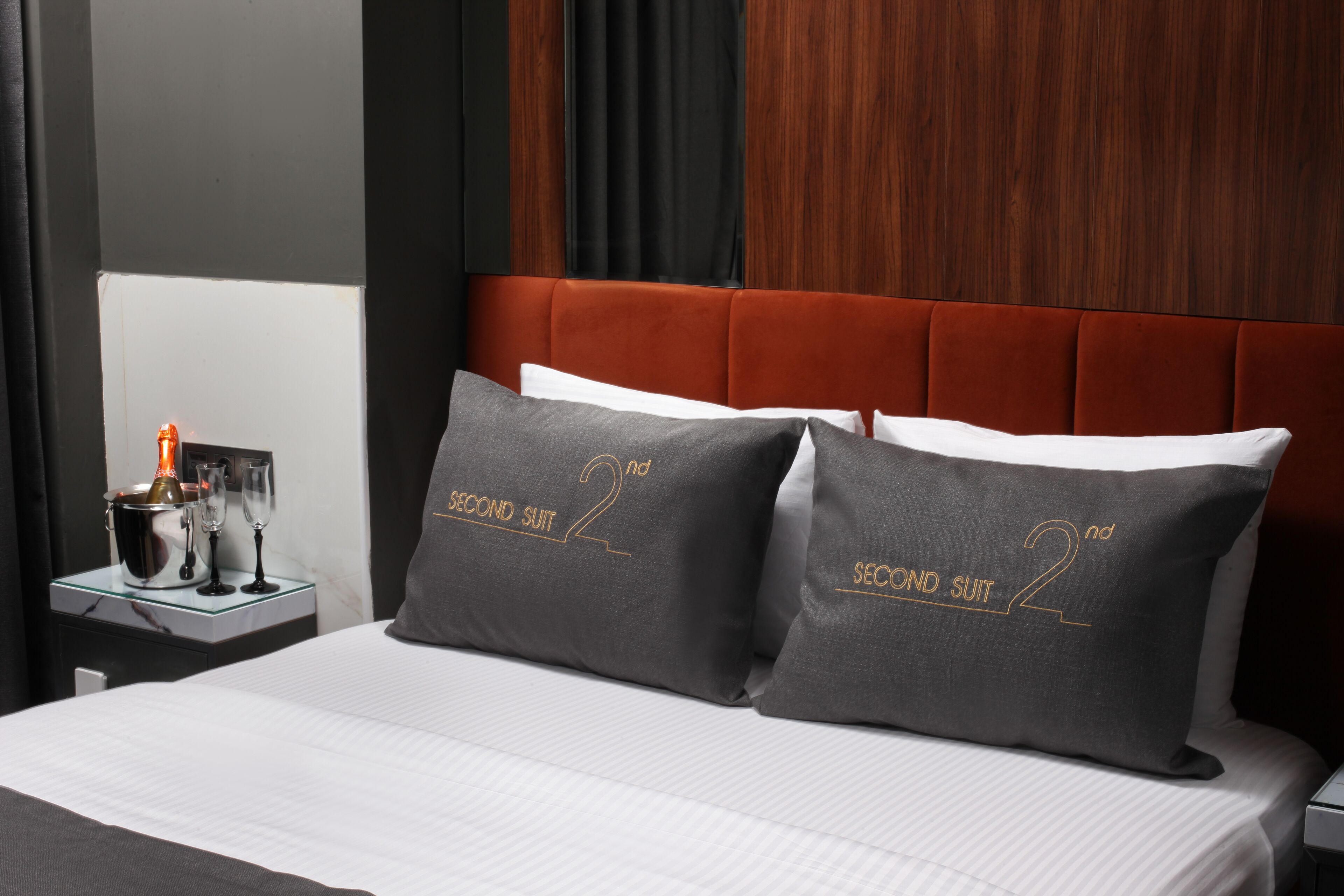 Una habitación de hotel con una cama, dos almohadas con el texto "SECOND SUIT", una mesita de noche con una vela y vasos, y un cabecero de madera.