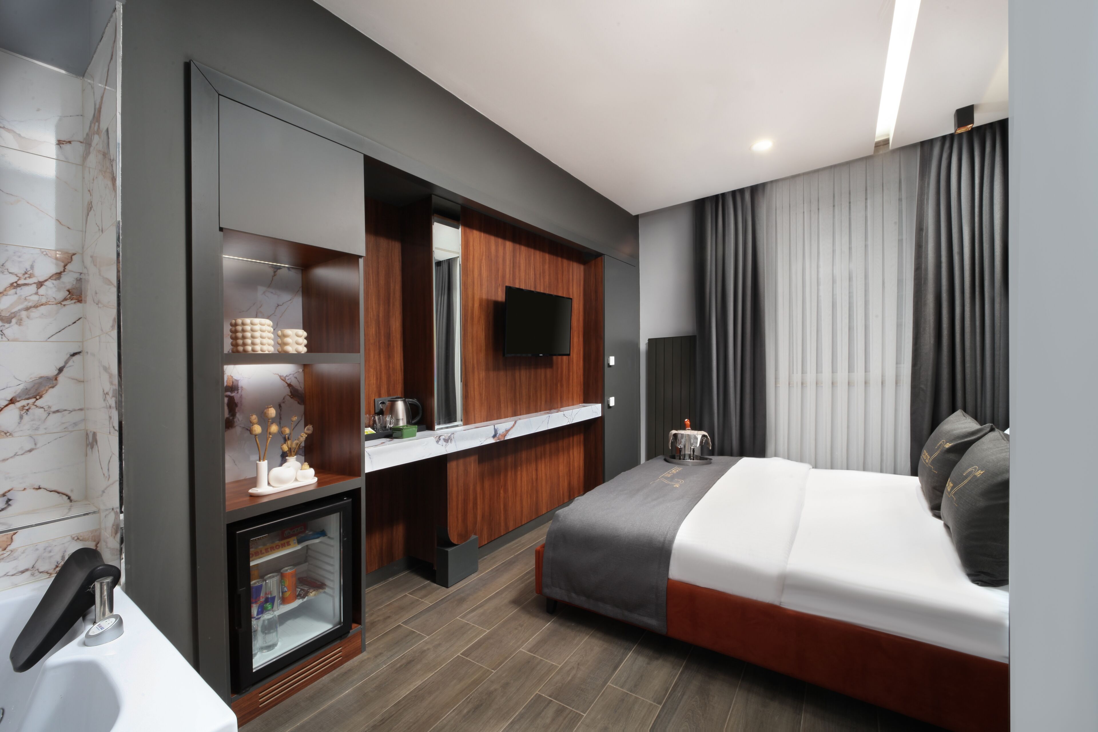A modern hotel room with a bed, a built-in TV, a minibar, and a bathroom with a marble wall.