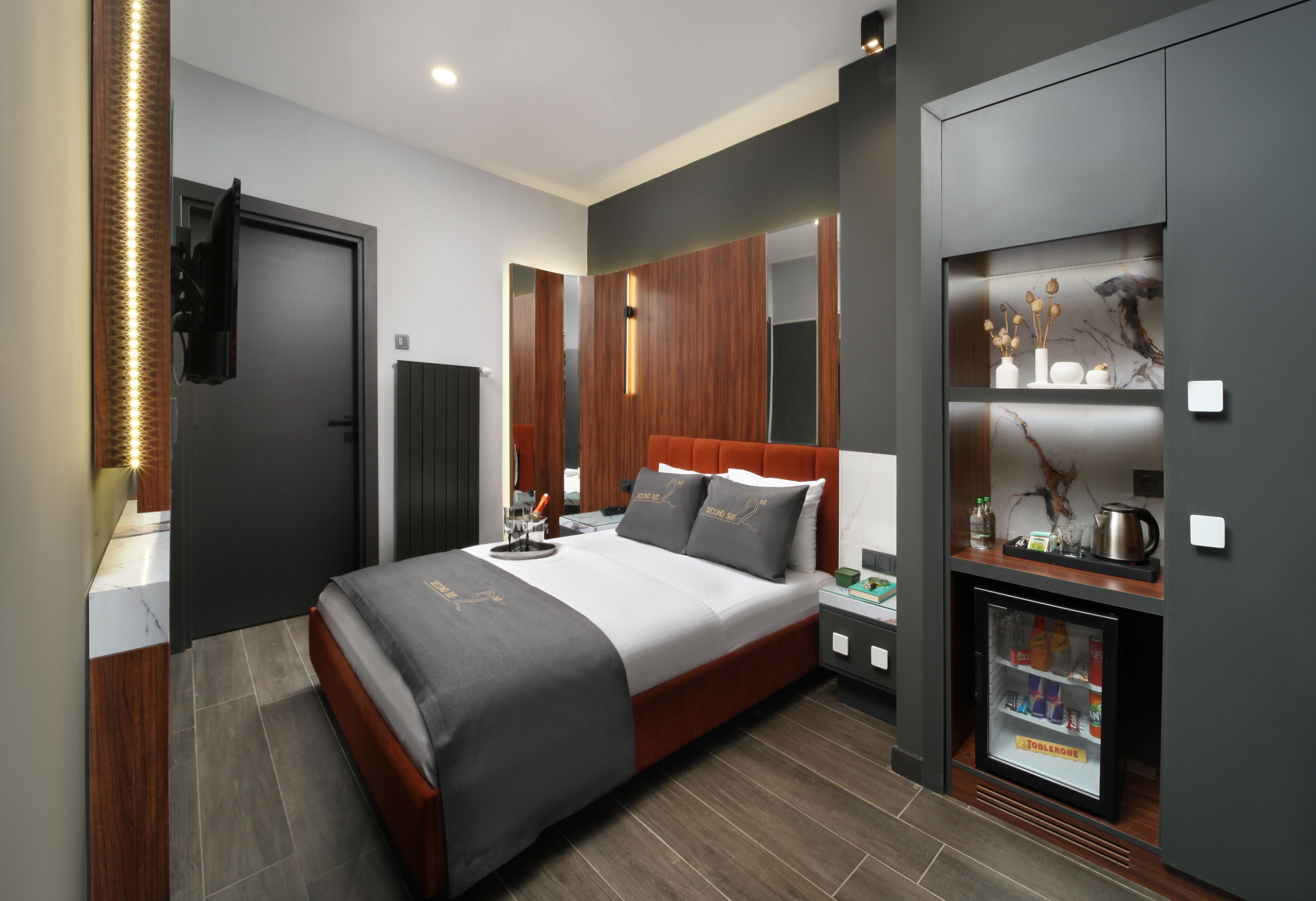 A modern hotel room with a large bed, built-in wardrobe, bedside tables, a minibar, and a TV.