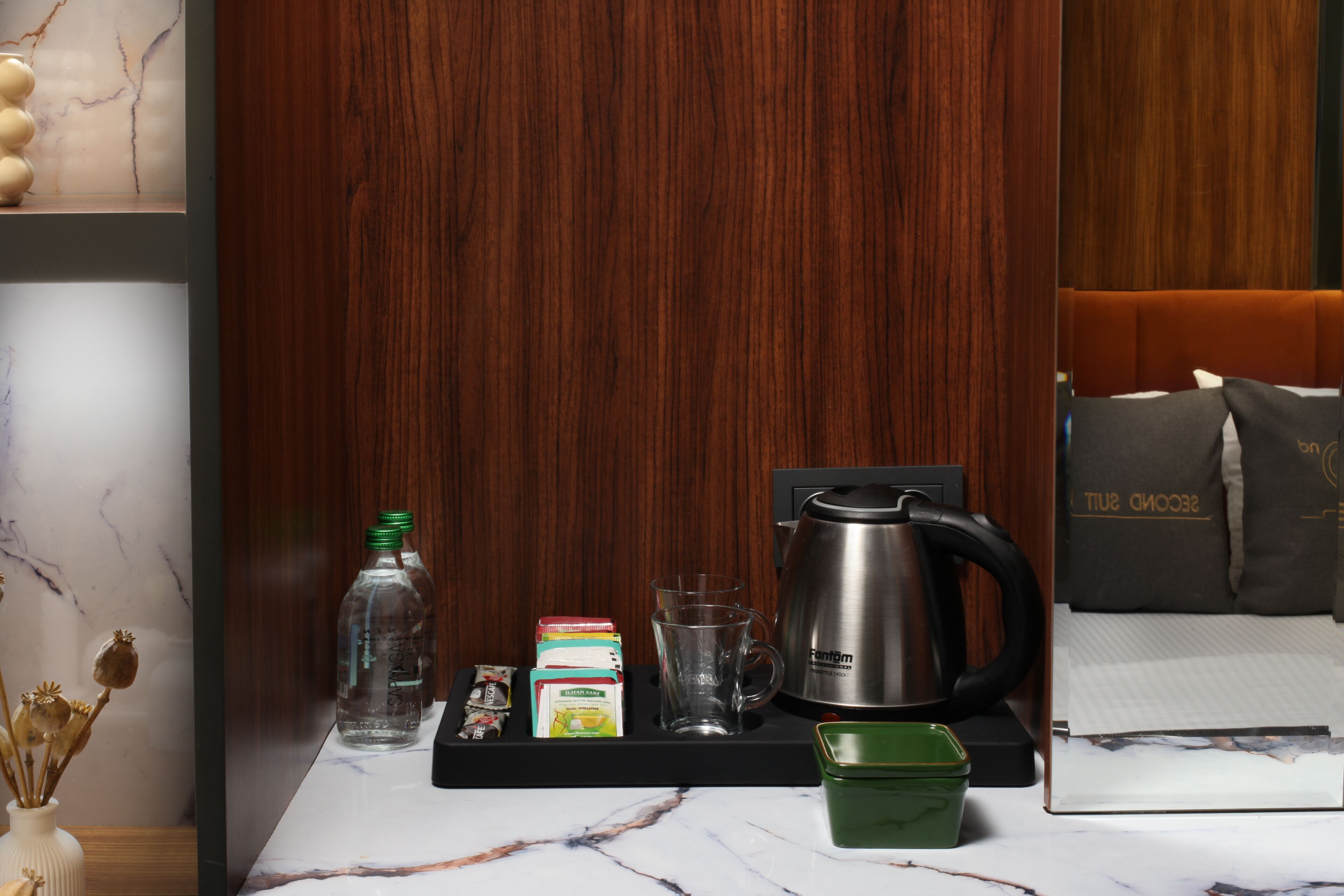 Hotelkamer met een wand van houten panelen, een tafel met marmeren blad waarop een dienblad met een waterkoker, een glas, een fles en ontbijtartikelen staan.