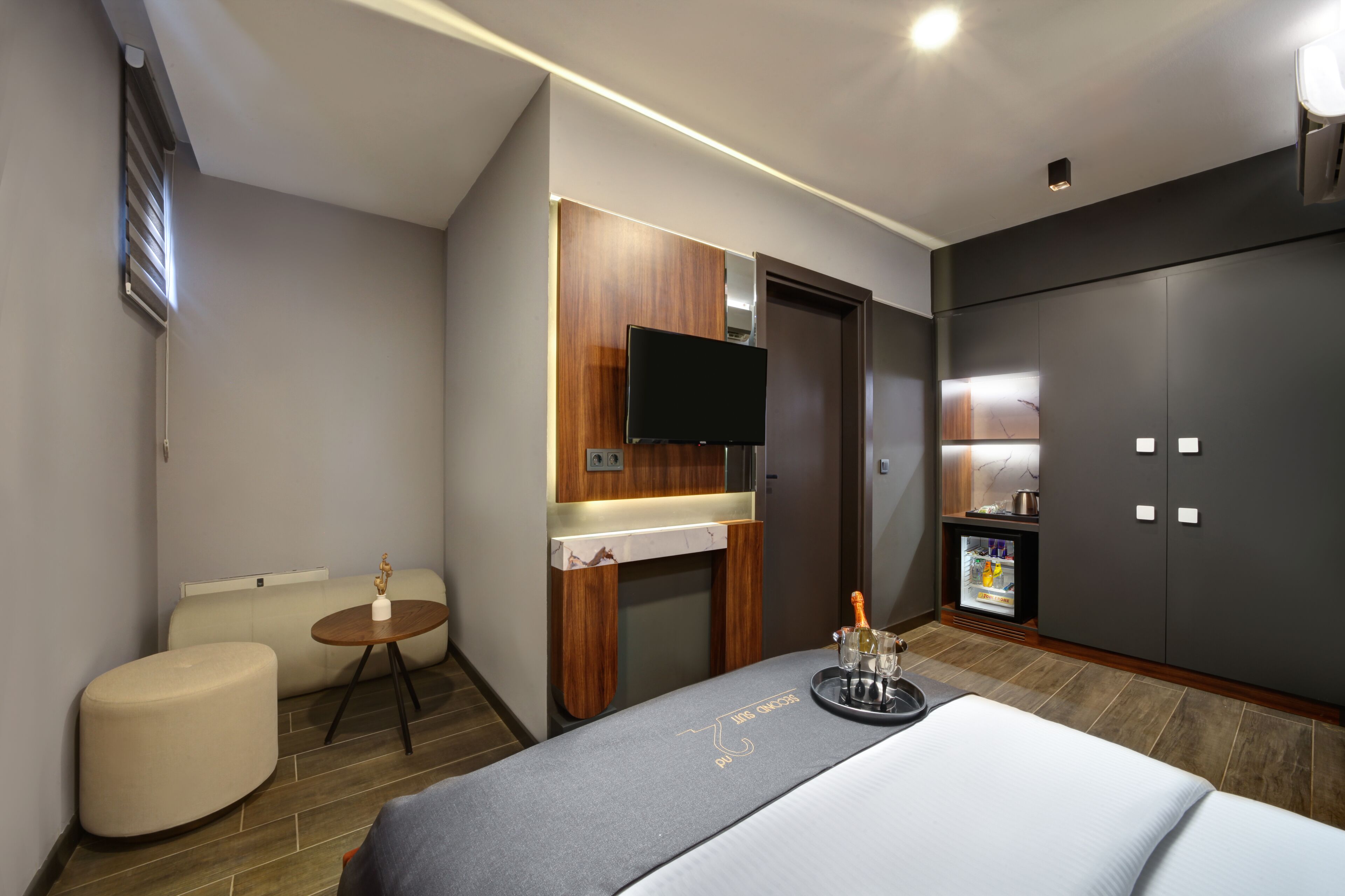 A modern hotel room with a bed, a small round table with a bottle, a TV mounted on the wall, and a minibar.