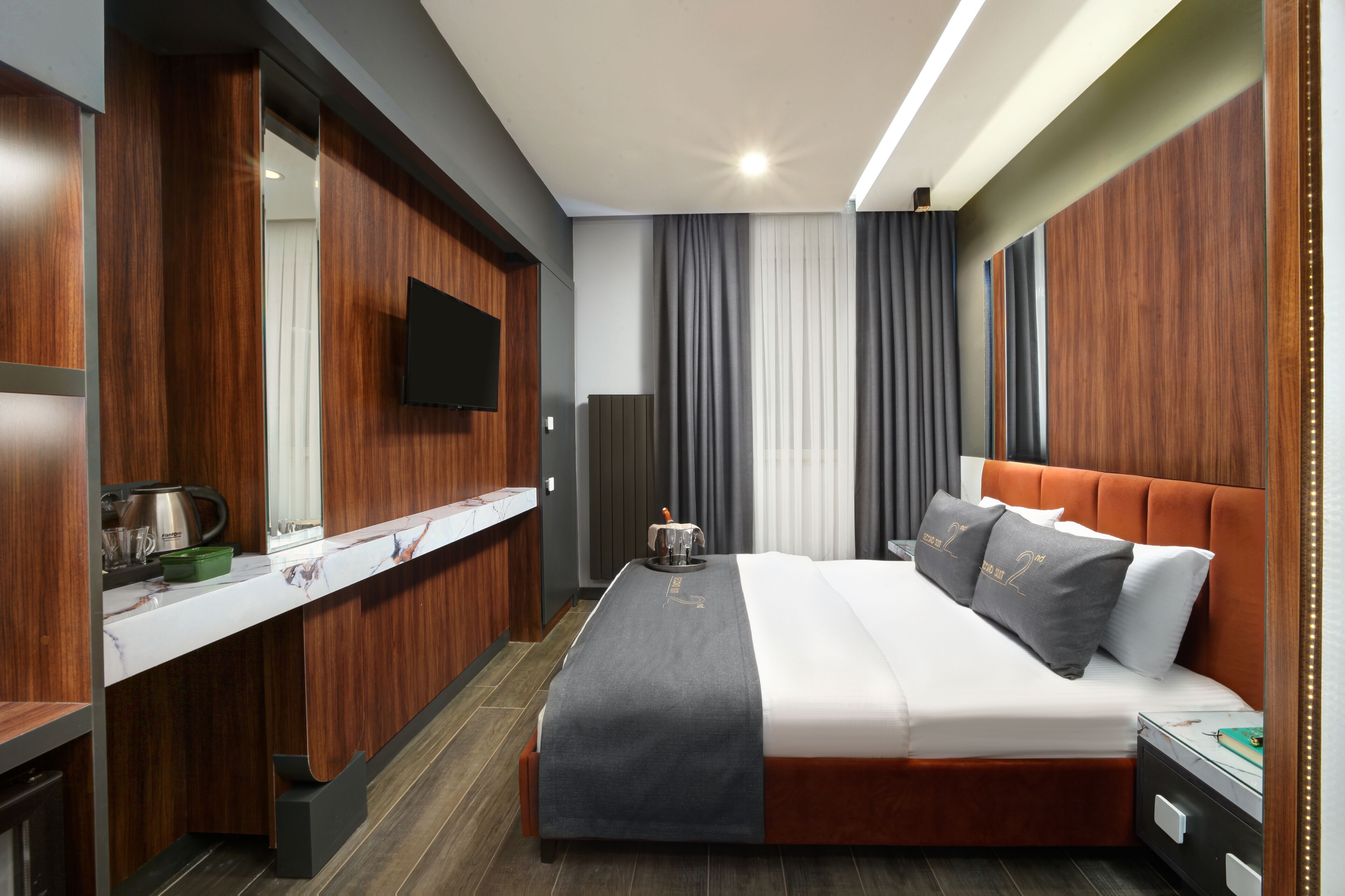 Ein modernes Hotelzimmer mit einem hölzernen Schminktisch, einem Flachbildfernseher, einem Bett mit grau-weißer Bettwäsche und einem Nachttisch.