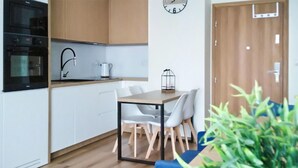 Cozinha moderna com armários brancos, forno preto e bancada de madeira. Há uma mesa de jantar com cadeiras brancas e almofada azul. Um relógio está na parede, e uma porta com puxador é visível.
