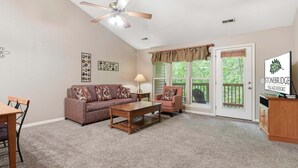 A living room with a sofa, armchair, coffee table, and a dining table. There is a ceiling fan, a lamp, and a sign for Stonebridge Village Resort. - Golf Condo + Connecting Suite at StoneBridge Resort (Reeds Spring)