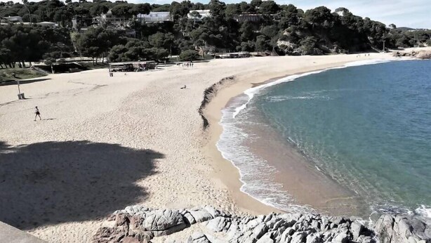 Een zandstrand met helderblauw water, een rotsachtige kustlijn en een beboste heuvel op de achtergrond.