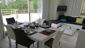Een moderne woonkamer met een eethoek. Er staat een witte eettafel met zwarte stoelen, een blauwe kussen op een bank en een televisie aan de muur.