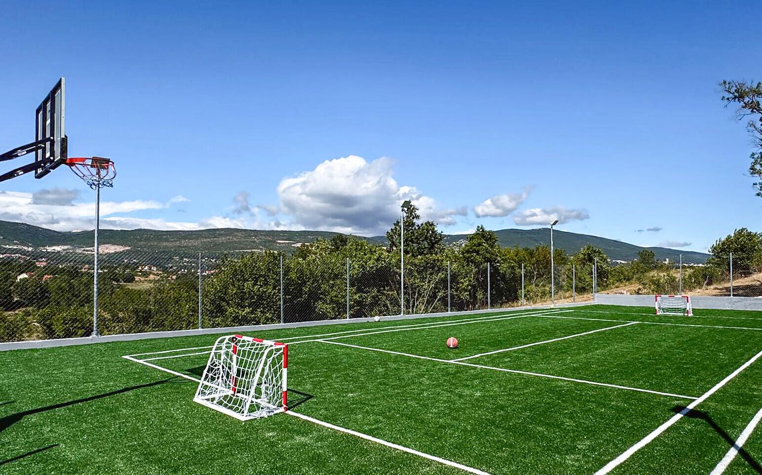 Eine Sportanlage im Freien mit Basketballkorb und Fußballtor, umzäunt und von Bäumen gesäumt, mit Blick auf ferne Hügel.