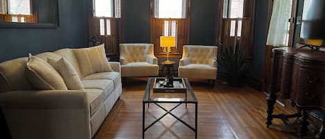 Een woonkamer met een bank, twee fauteuils, een salontafel, een kroonluchter en houten vloeren.