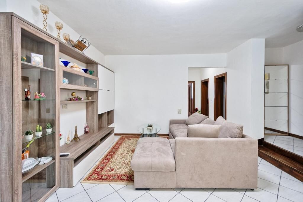 A living room with a beige sofa, a glass coffee table, a wooden bookshelf, and a red patterned rug.
