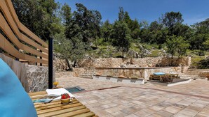 Un patio con muros de piedra, mobiliario de madera y vistas a árboles y colinas.