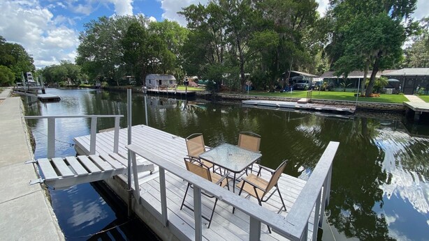 Un muelle con una mesa y sillas, una masa de agua tranquila y vegetación circundante.