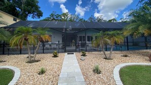 Una casa moderna con un porche cubierto, una piscina y un jardín bien cuidado con palmeras y guijarros.