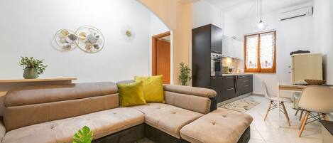 Een moderne woonkamer met een hoekbank, een eethoek met tafel en stoelen, en een keuken op de achtergrond.