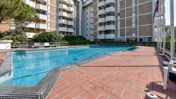 Une piscine dotée d’une terrasse carrelée, entourée d’un immeuble à plusieurs étages avec des balcons.