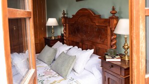 Een slaapkamer met een houten hoofdbord, twee nachtkastjes met lampen, een bed met veel kussens, en een deur die naar een andere kamer leidt.