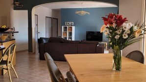 A dining area with a wooden table and chairs, a vase of flowers, a sofa, and a television in the background.